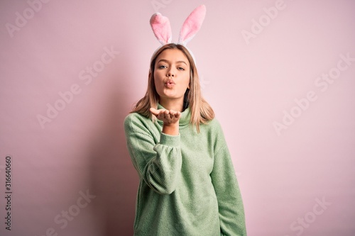 Young beautiful woman wearing easter rabbit ears standing over isolated pink background looking at the camera blowing a kiss with hand on air being lovely and sexy. Love expression.