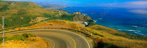 This is Route 1 also known as the Pacific Coast Highway. The road curves around a bend to the left and drops down overlooking the ocean. The rocky hillside is also seen next to the ocean.