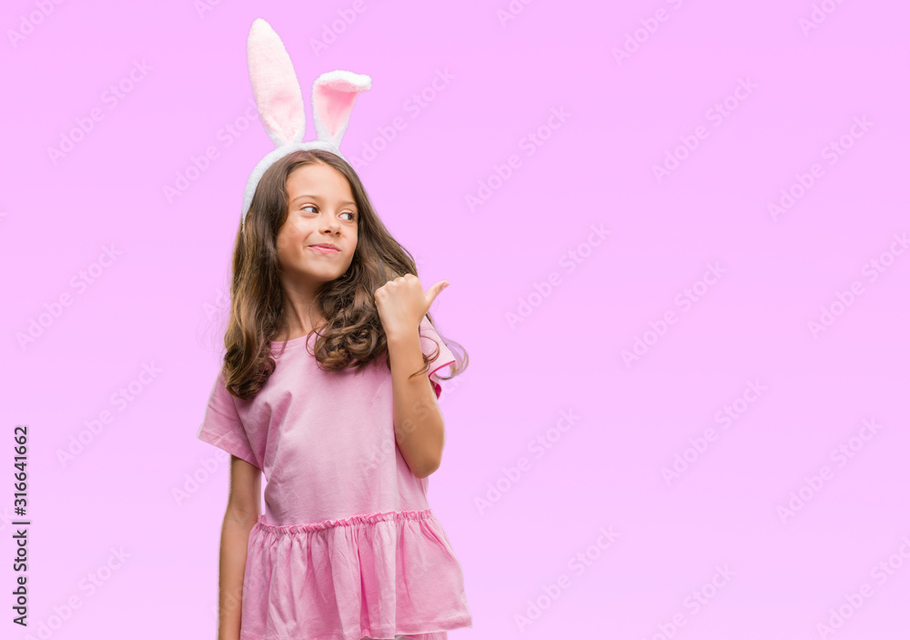 Brunette hispanic girl wearing easter rabbit ears pointing and showing with thumb up to the side with happy face smiling