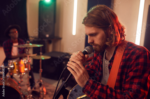Side view portrait of handsome bearded man singing to microphone during rehearsal or concert with music band in dimly lit studio, copy space