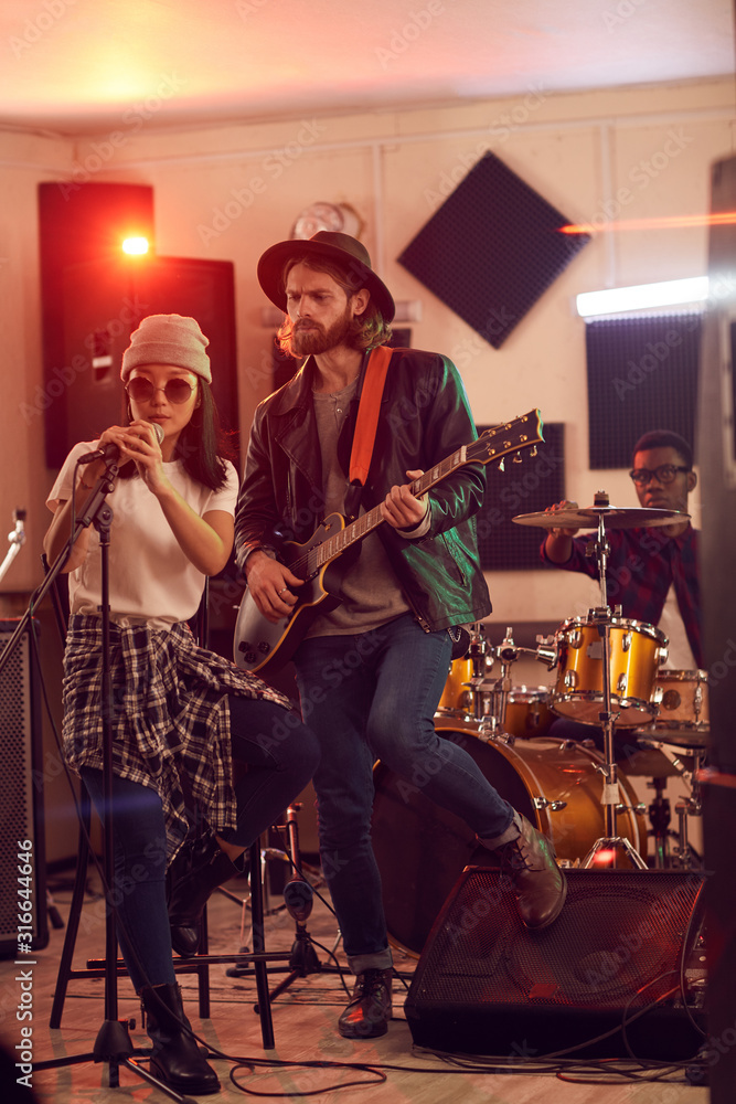 Full length portrait of contemporary band playing music during rehearsal or concert in dimly lit studio, focus on handsome man playing guitar in foreground, copy space Full length portrait of contemporary band playing music during rehearsal or concert in dimly lit studio, focus on handsome man playing guitar in foreground, copy space