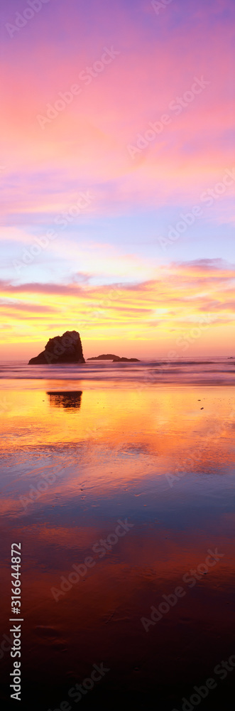 This is the Oregon coast at sunset. The large rock to the left is referred to as one of the sea stacks on the beach. The pink and blue sunset sky is reflected in the water on the beach.