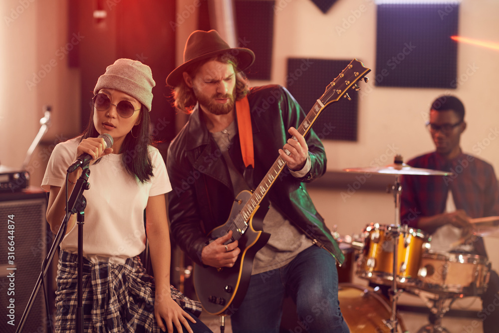 Fototapeta premium Portrait of contemporary band playing music during rehearsal or concert in dimly lit studio, focus on young Asian woman singing to microphone in foreground, copy space