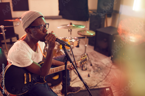High angle portrait of contemporary African-American man singing to microphone and playing guitar during rehearsal or concert with music band in recording studio, copy space photo