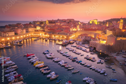 Aerial view of boats and yachts and beautiful architecture at night in Dubrovnik, Croatia. Top view of old city at sunset. Port in the sea, city lights, historical centre, buildings and colorful sky