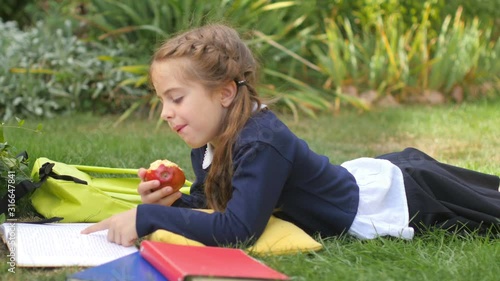 A schoolgirl is lying on the grass with an apple reading a book and studying alone in the park. School lunch. Schoolgirls learning in kid education concept