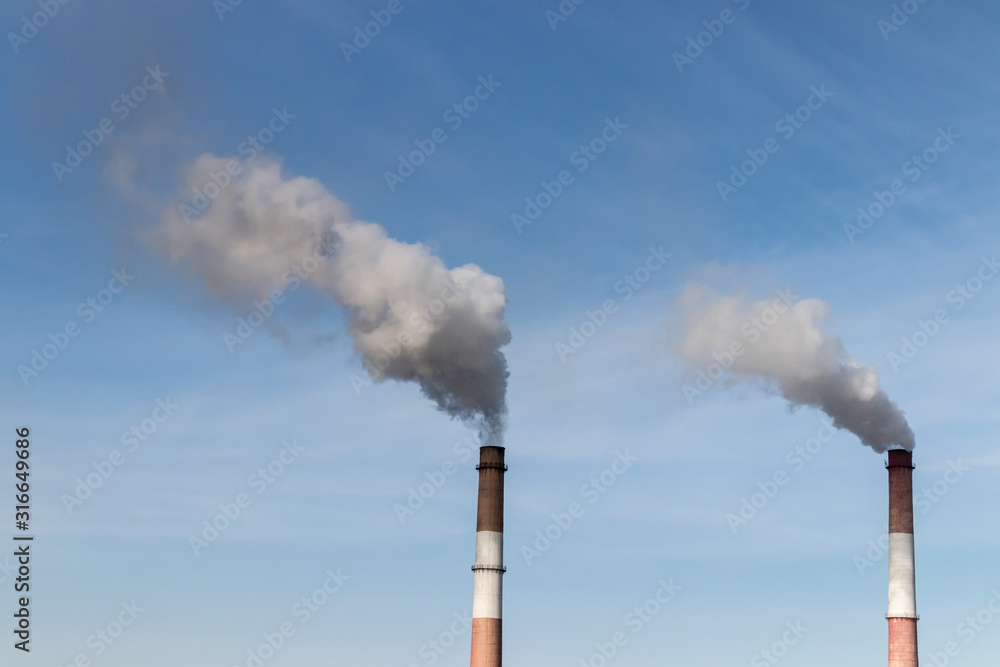 Two pipes of a thermal power plant release gray-white steam into the air against a blue sky Two pipes of a thermal power plant release gray-white steam into the air against a blue sky