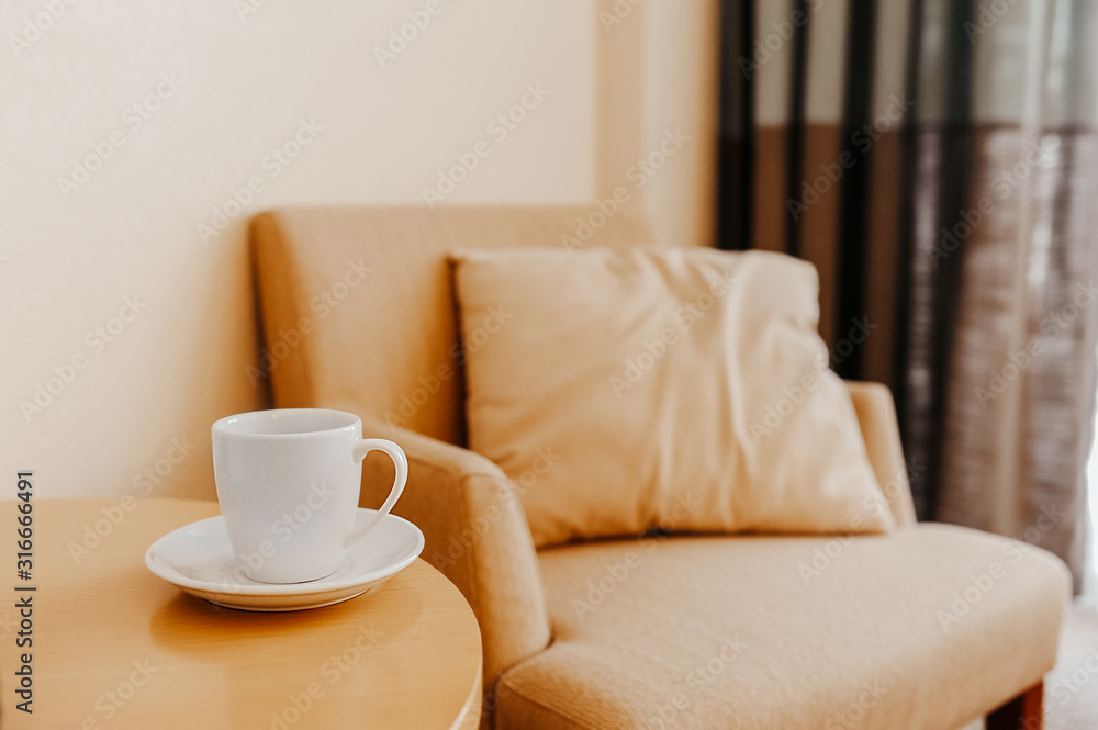 white cup on a wooden coffee table against the background of a beige armchair with a pillow, elegant interior in the style of minimalism, a place for relaxation or business meetings