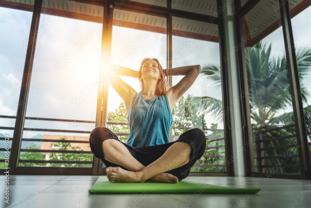 An attractive 40-year-old middle-aged woman practices yoga in a panoramic window room overlooking the garden at dawn in the sunshine. Meditation mindfulness healthy lifestyle concept - obrazy, fototapety, plakaty 