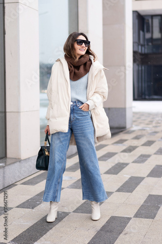 European model girl in a beige oversized down jacket, knitted sweater, flared jeans with a handbag and glasses, walks near the office building. Life style
