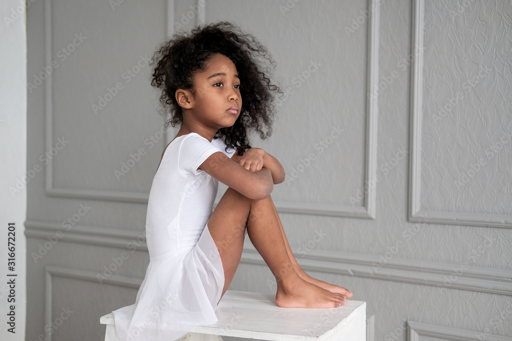 A portrait of an African ballerina in a white gymnastic costume sits on a wooden chair. A portrait of an African ballerina in a white gymnastic costume sits on a wooden chair.