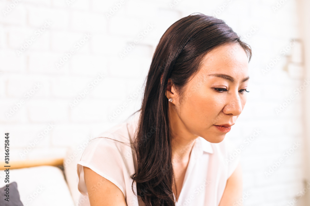 Side view of middle aged beautiful Asian woman sitting crying and wiping out tears and thinking about problems indoors Side view of middle aged beautiful Asian woman sitting crying and wiping out tears and thinking about problems indoors