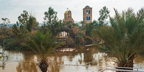 site of the baptism of jesus on the jordan river showing ancient churches in the background with palm trees and border markers in the foreground