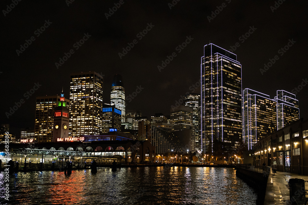 San Francisco port illuminated at night, against the backdrop of impressive skyscrapers with light and reflection in the water, view from the side of the San Francisco Bay.
