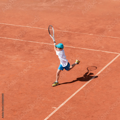 A boy plays tennis on a clay court. A little tennis player focused on the game and shot in flight after hitting the ball. Sports action frame. Active games. Square size. © Elena