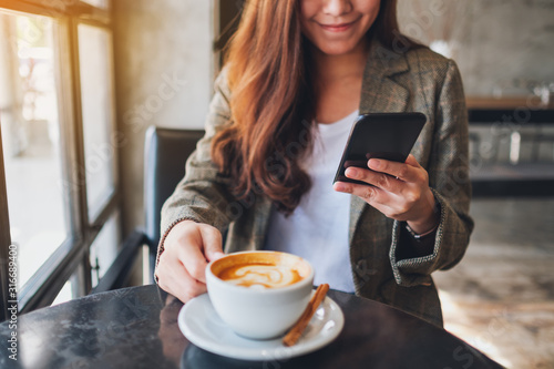 Closeup image of an asian woman holding and using mobile phone while drinking coffee in cafe