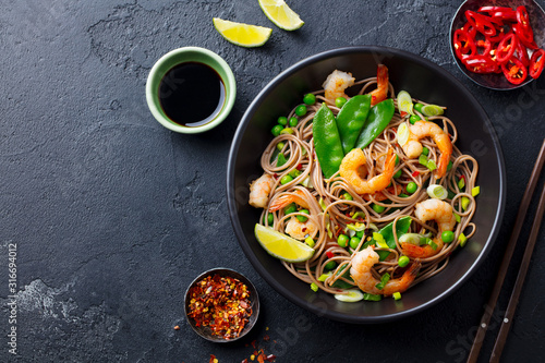 Stir fry noodles with vegetables and shrimps in black bowl. Slate background. Top view. Copy space.