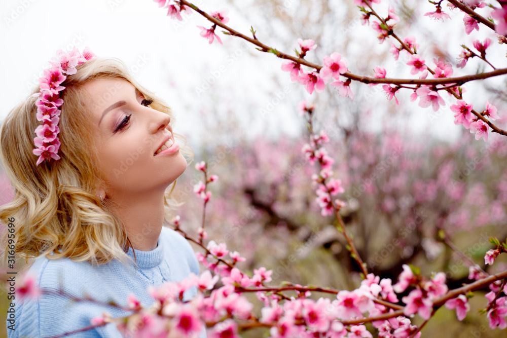 Fototapeta premium Portrait of beautiful woman posing against the pink flower trees garden in spring, dressed blue sweater and wreath pink flower