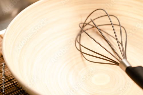 Selective focus, gray wire of modern design balloon whisk in clean and neat natural round bamboo mixing bowl on wooden plate mat on cooking counter in kitchen with warm morning sunlight shine through.