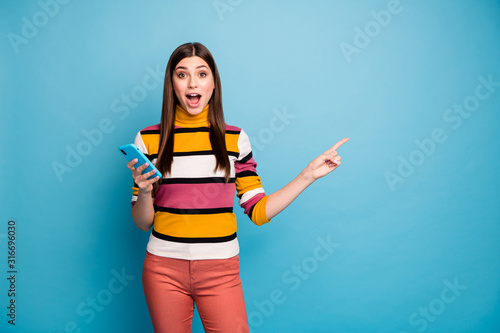 Astonished crazy girl point index finger copyspace use smart phone indicate incredible ads promotion impressed scream wow omg wear red pants trousers jumper isolated blue color background