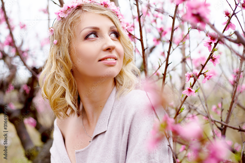 Fototapeta premium portret pięknej kobiety w ciąży pozowanie na tle różowych drzew kwiatowych ogród i mgła wiosną, ubrany kardigan w proszku i wieniec różowy kwiat. Kobieta trzyma brzuch.