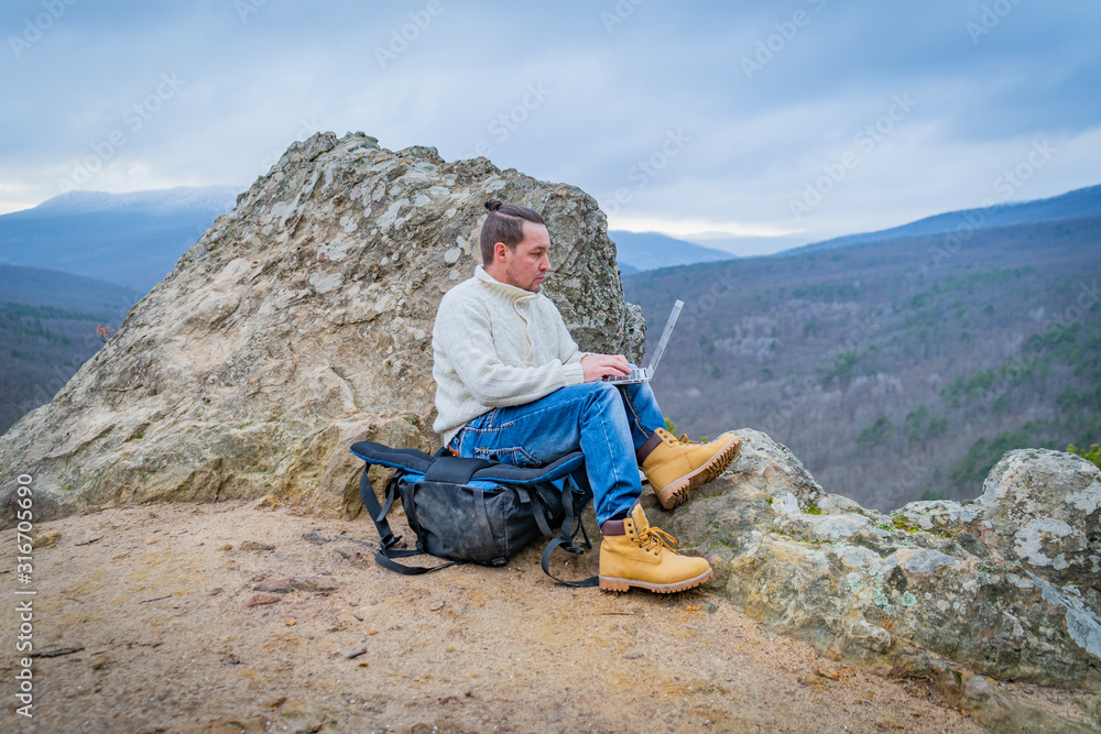 Well-dressed man working with laptop sitting on the rocky mountain on beautiful scenic clif background