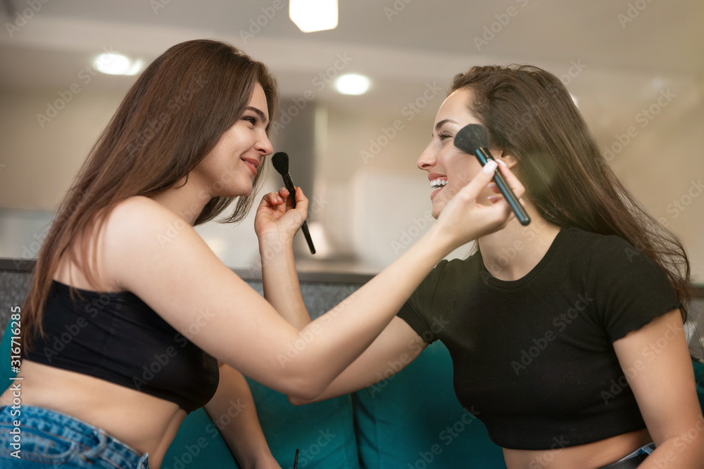 two young beautiful women friends sitting on the sofa getting ready for party doing makeup , applying each other blush with makeup brushes , beauty day routine two young beautiful women friends sitting on the sofa getting ready for party doing makeup , applying each other blush with makeup brushes , beauty day routine