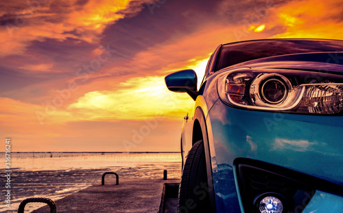 Blue compact SUV car with sport and modern design parked on concrete road by the sea beach at sunset. Front view of luxury car. Closeup SUV fog lamp and headlights with beautiful sunset sky in summer. © Artinun