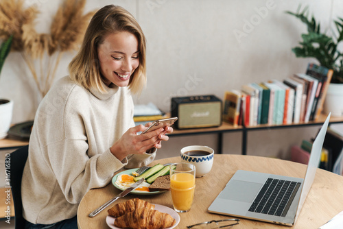 Photo of woman using cellphone and laptop while having breakfast at home