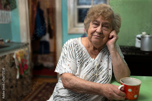 Sad elderly woman sits at a table at home in the kitchen