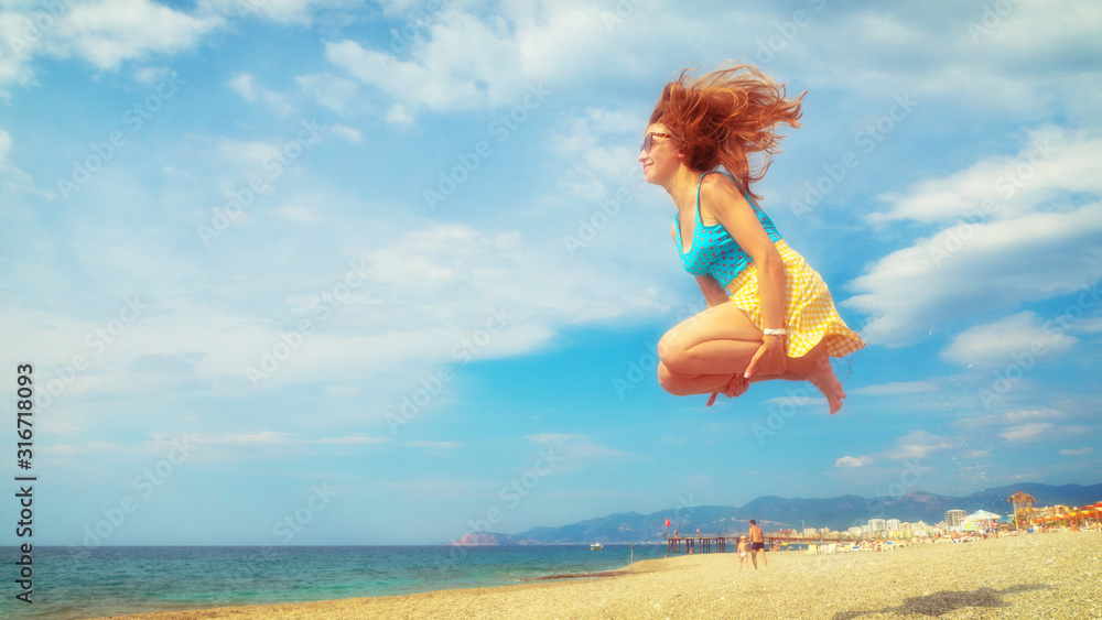 Naklejka premium Excited young woman having fun on beach in summer. Side view of carefree youthful female tourist in sunglasses enjoying freedom and jumping on sandy seashore