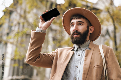 Image of stylish young man holding cellphone while walking outdoors