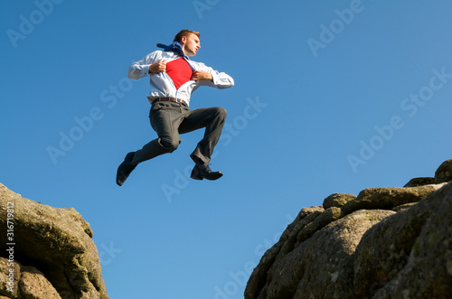 Superhero businessman taking a daring leap between two boulders outdoors against bright blue sky