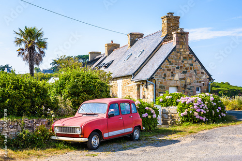 A vintage french car parked in front of a typical granite breton house with slate roof, palm tree and hydrangea by a sunny summer day in Brittany, France.