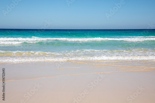 Cloudless sky over a colorful sea with diferent colors aquamarine turquoise dakr blue azure with waves prducing white foam crushing against a yellow beach summer sumertime sunny calm water tranquil © Markus Schmid