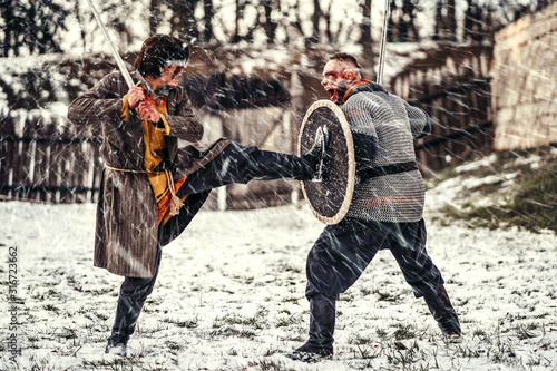 Two ancient warriors in armor with weapons fighting with swords in the snow