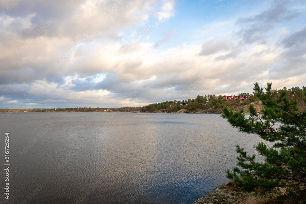 Panoramic view of Baltic Sea bay in spring. Landscape with beautiful green pines branches in front. Horizon, skyline. Islands with pines and houses. Scandinavian archipelago, Sweden. Panoramic view of Baltic Sea bay in spring. Landscape with beautiful green pines branches in front. Horizon, skyline. Islands with pines and houses. Scandinavian archipelago, Sweden.