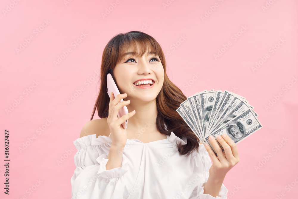 Happy Shocked ginger woman talking by smartphone while holding money and looking at the camera over pink background Happy Shocked ginger woman talking by smartphone while holding money and looking at the camera over pink background