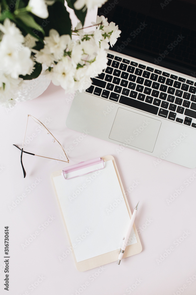 Office desk workspace with laptop, flowers bouquet and blank paper copy space mockup clipboard on pink table. Flat lat, top view freelancer co-working background. Office desk workspace with laptop, flowers bouquet and blank paper copy space mockup clipboard on pink table. Flat lat, top view freelancer co-working background.