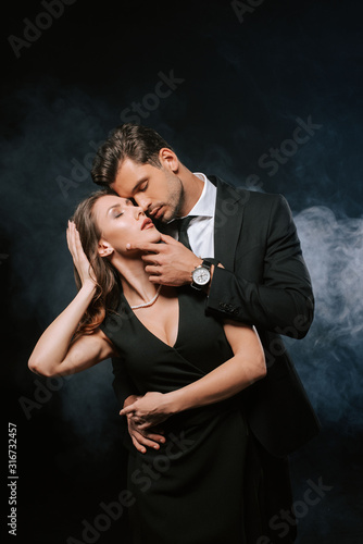 sensual man touching attractive woman in dress on black with smoke