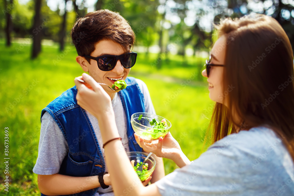 Fototapeta premium Young couple enjoying lunch outdoor on sunny day. Happy young friends eating natural vegetable salad for lunch. Healthy lifestyle. Concept picnic. Clean eating, vegan food concept.