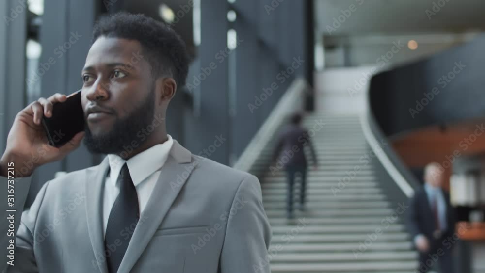 Chest-up tracking shot of confident young Afro-American man in business suit and tie walking through lobby of busy office building, talking on smartphone and smiling
