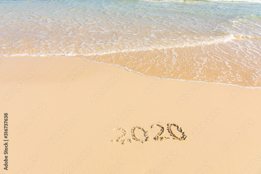 Fototapeta premium Summer beach holiday 2020 season golden sand - year - decade - new season lettering on the beach with wave and clear blue sea. Numbers 2020 year on the sea shore, message handwritten New Years concept
