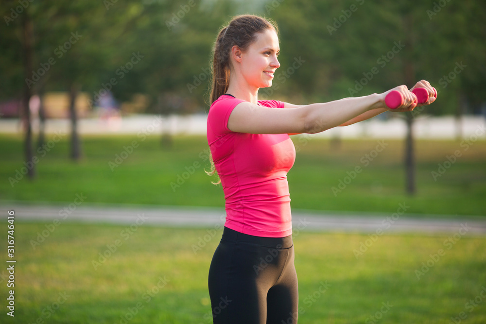 beautiful young woman in sportswear is engaged in training with dumbbells in the summer in the park beautiful young woman in sportswear is engaged in training with dumbbells in the summer in the park