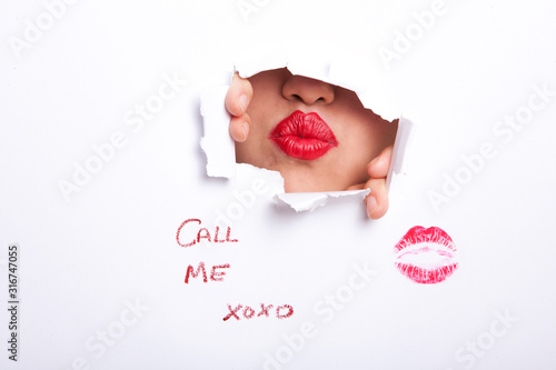 Tablou pe pânză Young woman's red puckered lips seen through ripped paper with text and lipstick