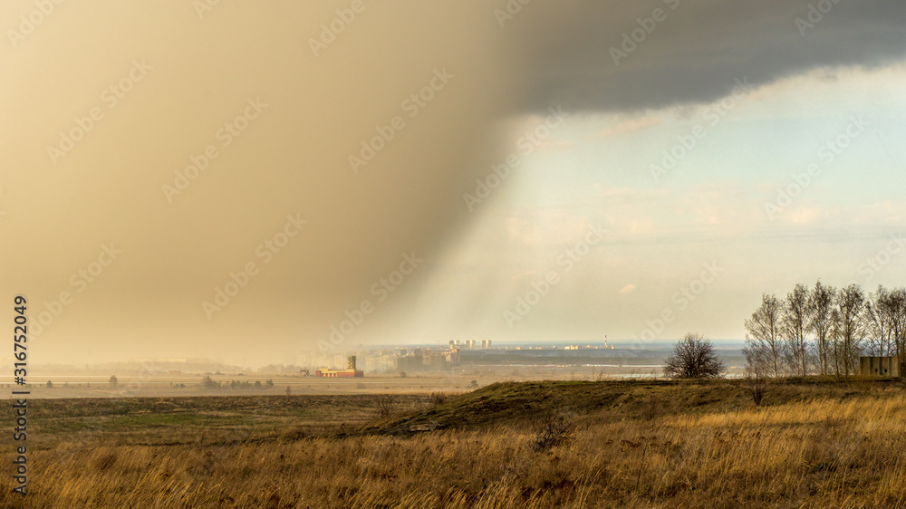 Spring natural and urban panoramic landscape with approaching huge storm cloud and heavy rain over burnt field and large city on horizon
