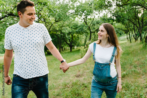 Pregnant girl and her husband are happy to hold hands,  goes in the outdoor in the garden background with trees. Close up. upper half. Look at each other. © Serhii