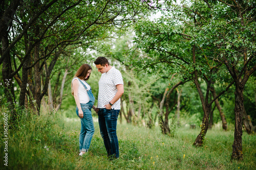 Pregnant girl and her husband are happy to hold hands, stand in the outdoor in the garden background with trees. full length. Looking down at the stomach.