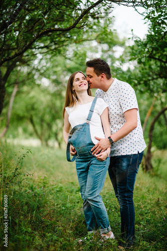 Pregnant girl and her husband kiss are happy to hug, hold hands on stomach, stand on grass in the outdoor in the garden background with trees. Close up. full length. Looking up.