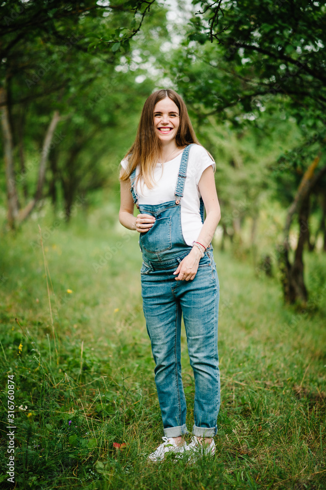 Pregnant happy girl stand and hold hands on stomach, stand on grass in the outdoor in the garden background with trees. Close up. full length. Looking at camera Pregnant happy girl stand and hold hands on stomach, stand on grass in the outdoor in the garden background with trees. Close up. full length. Looking at camera